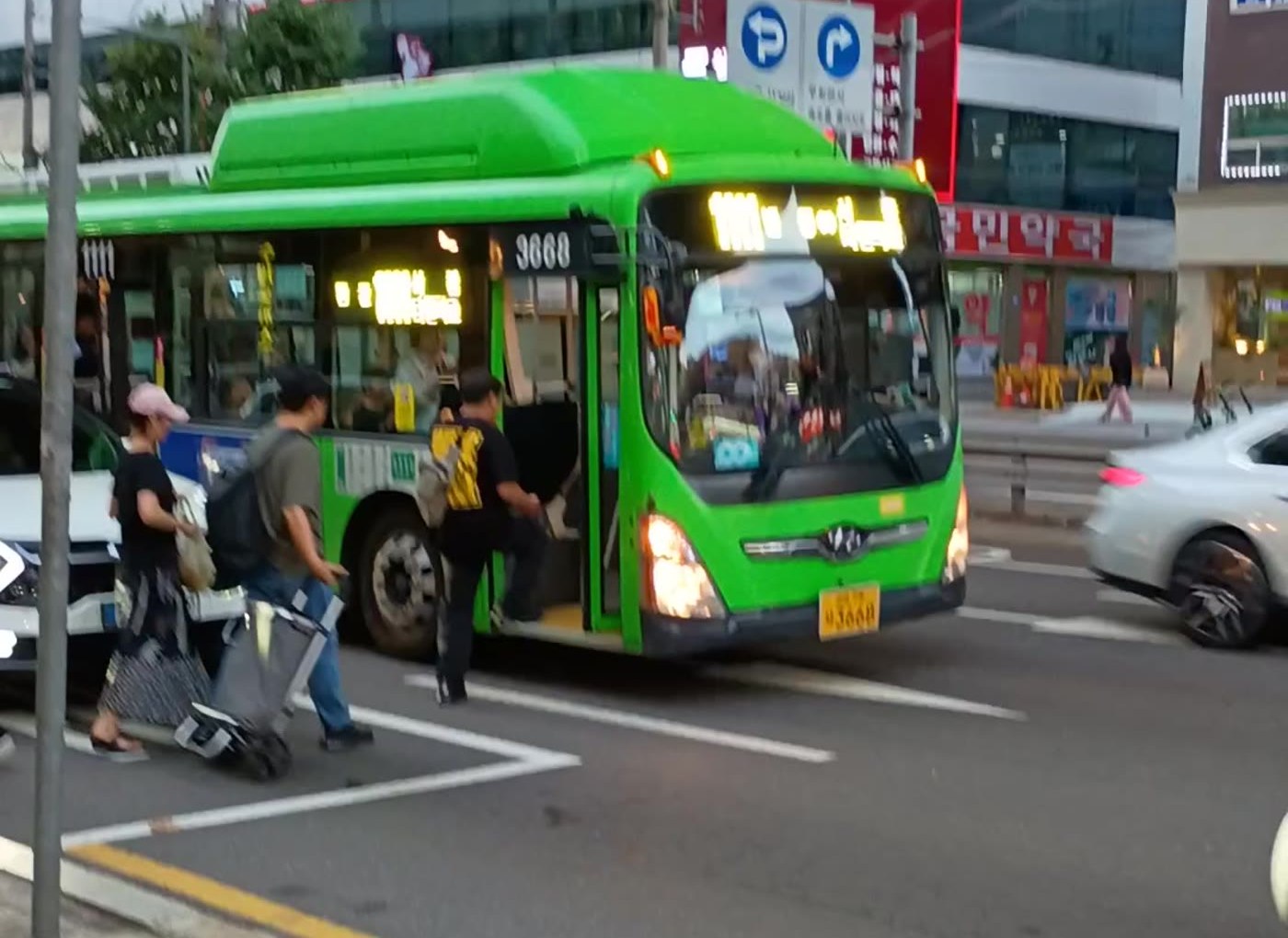 정류장 앞 연석에 흰색 승용차가 서 있고 녹색 버스는 차로에 정차, 승객들이 차도에서 앞문 계단을 오르는 장면