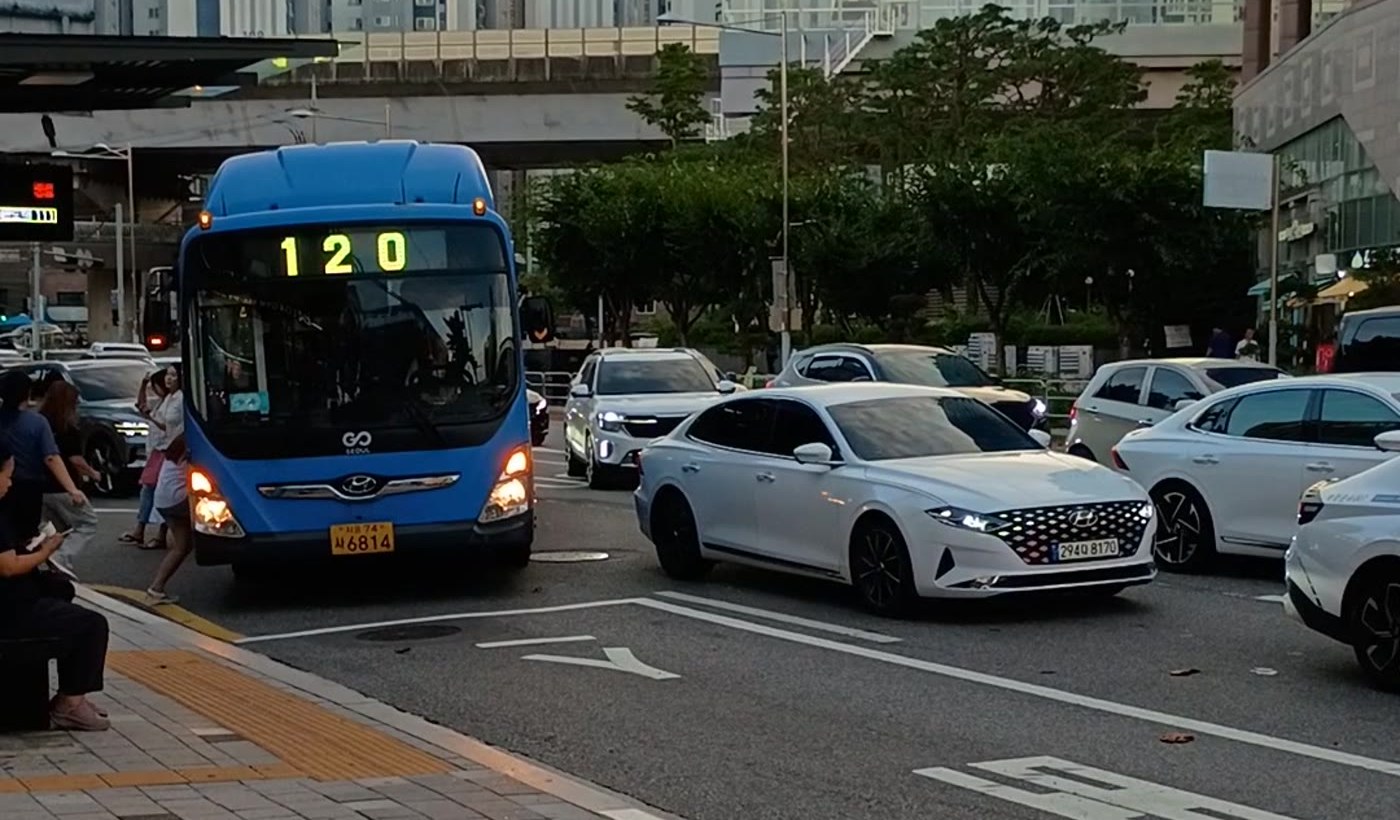 차로에 정차한 파란 120번 버스, 버스 앞 차도를 걸어 보도로 향하는 사람, 옆 차로를 지나가는 승용차 행렬